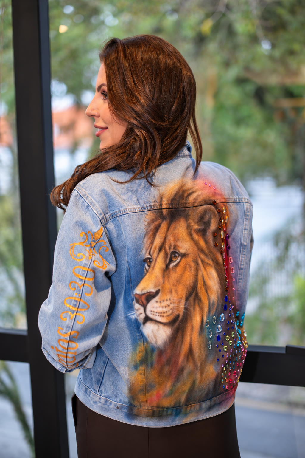 Mulher de perfil vestindo jaqueta jeans com leão pintado à mão, cascata de pedras coloridas e versículo dourado na manga