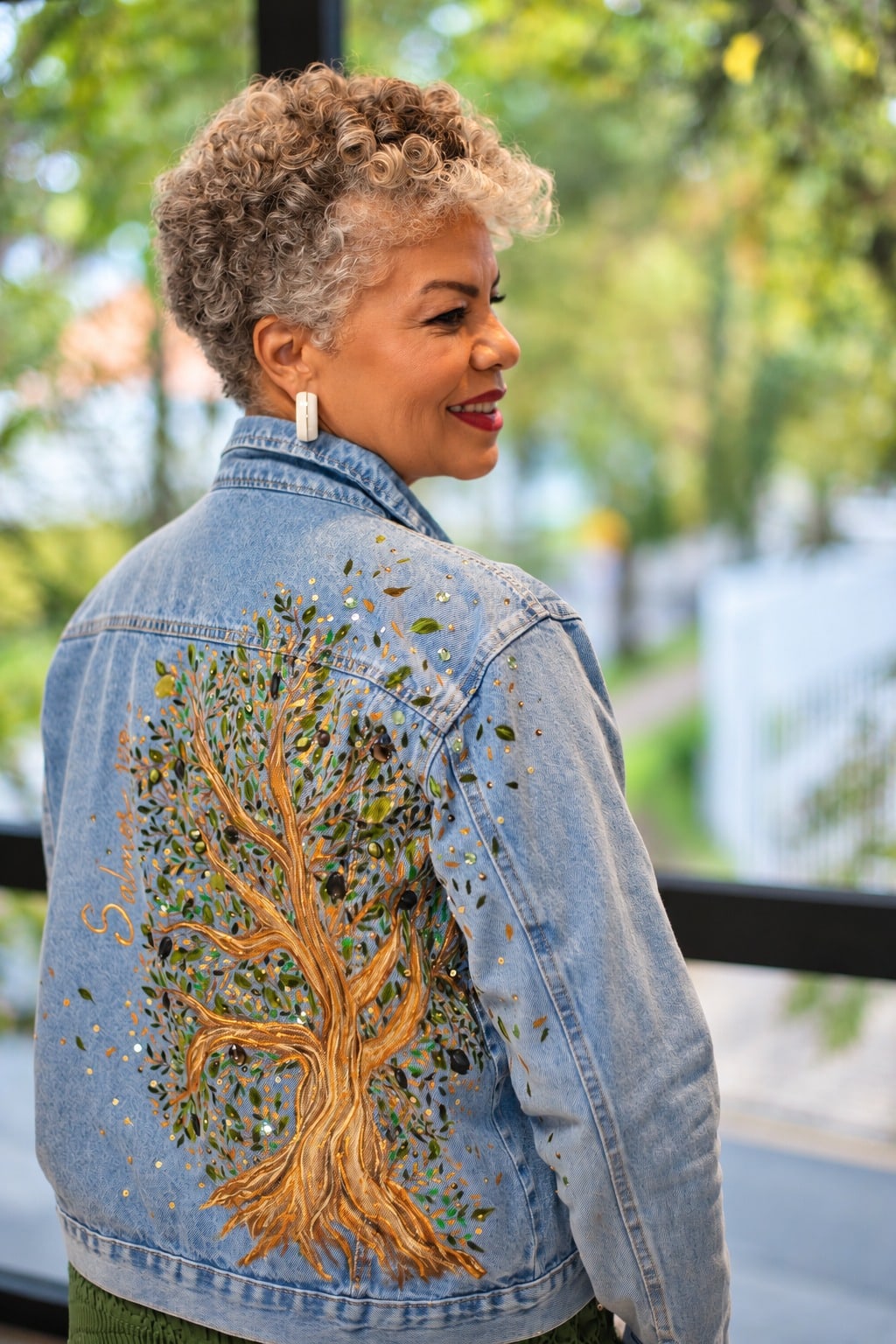 Mulher de cabelo grisalho cacheado de costas, sorrindo de perfil, com a jaqueta Oliveira: árvore de tronco dourado pintada à mão, folhas bordadas e brilhos