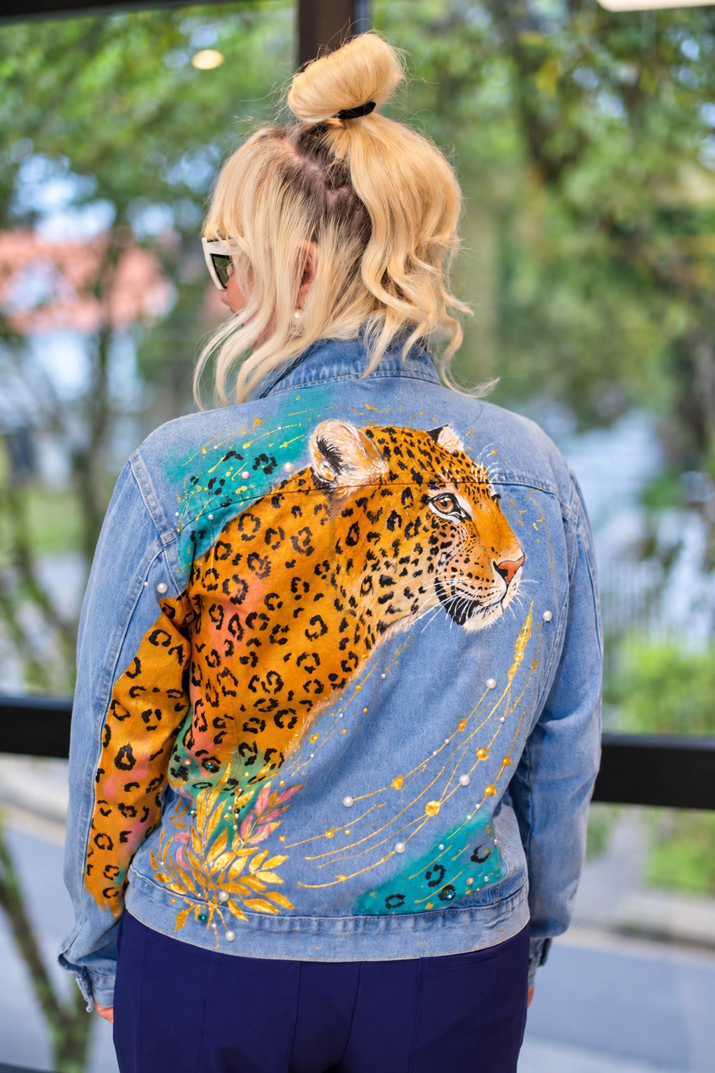 Mulher loira de costas vestindo jaqueta jeans com onça-pintada pintada à mão, folhas douradas, pérolas e traços em dourado